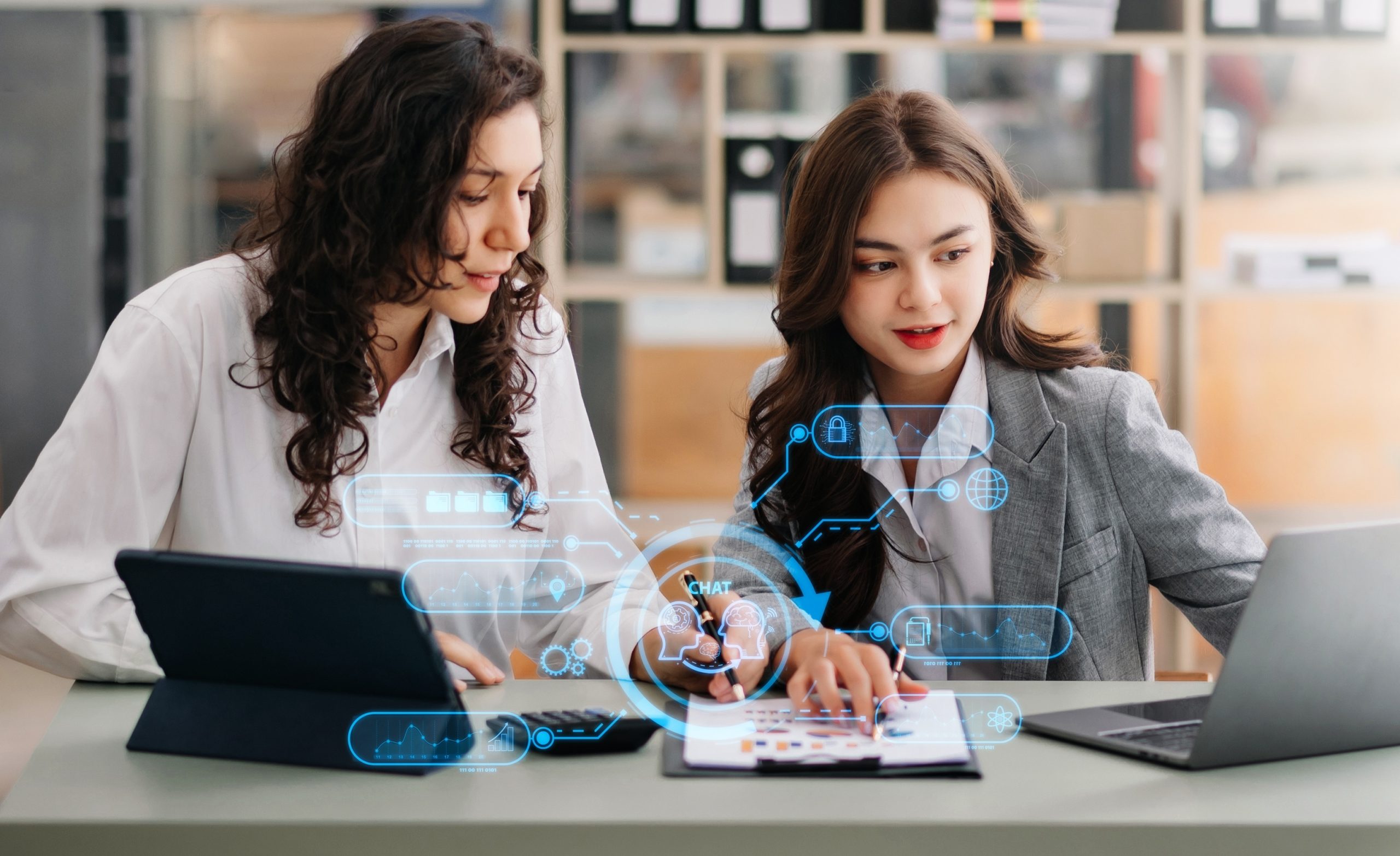 multifamily ai tools Two women at a table using digital devices, with holographic icons appearing between them. mpro Digital Edge
